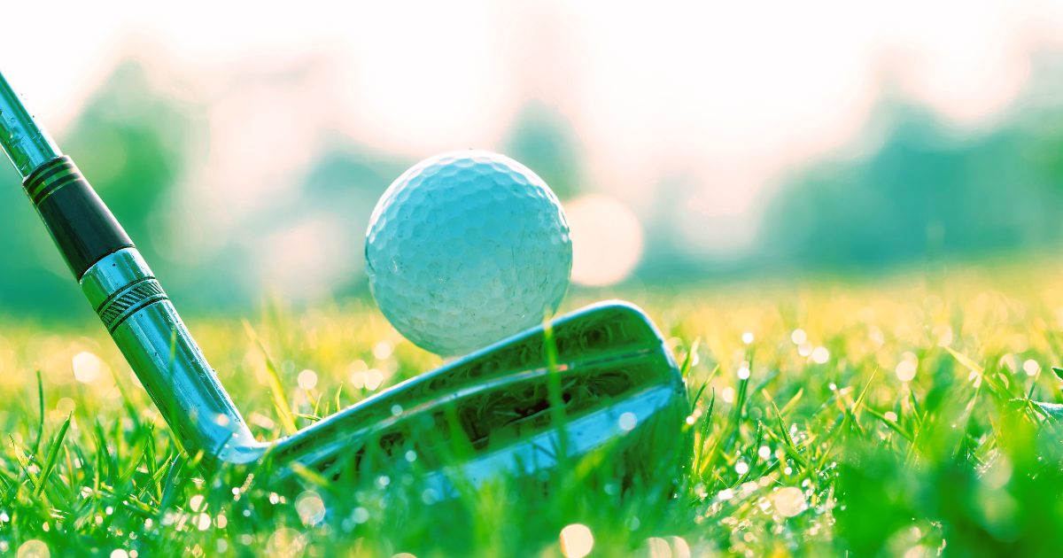 A close-up shot of a golf ball lying on grass with its club ready for use is shown, creating an early morning atmosphere on a golf course. Sunlight creates a gleaming effect in this scene that recalls early morning golf course activities.